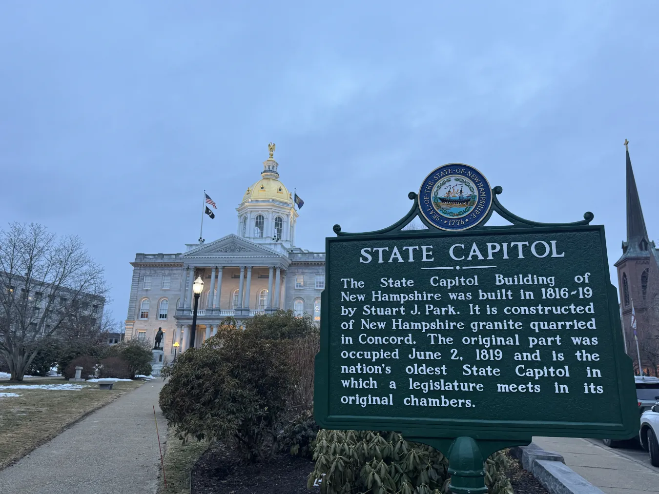 The New Hampshire State Capitol historical sign in the foreground at dusk; the New Hampshire State House in the background.