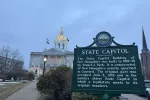 The New Hampshire State Capitol historical sign in the foreground at dusk; the New Hampshire State House in the background.