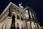 An image of the New Hampshire State House at night, with the Christa McAuliffe statue in the foreground.
