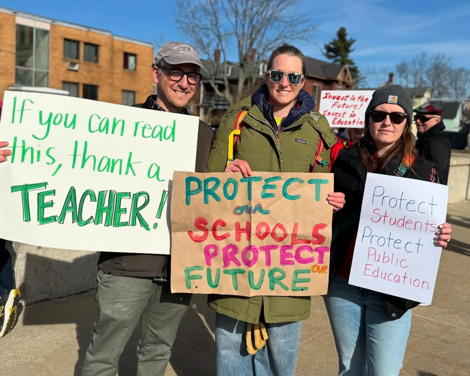 Members stand with signs that say "If you can read this thank a teacher," "Protect our Schools. Protect our Future."