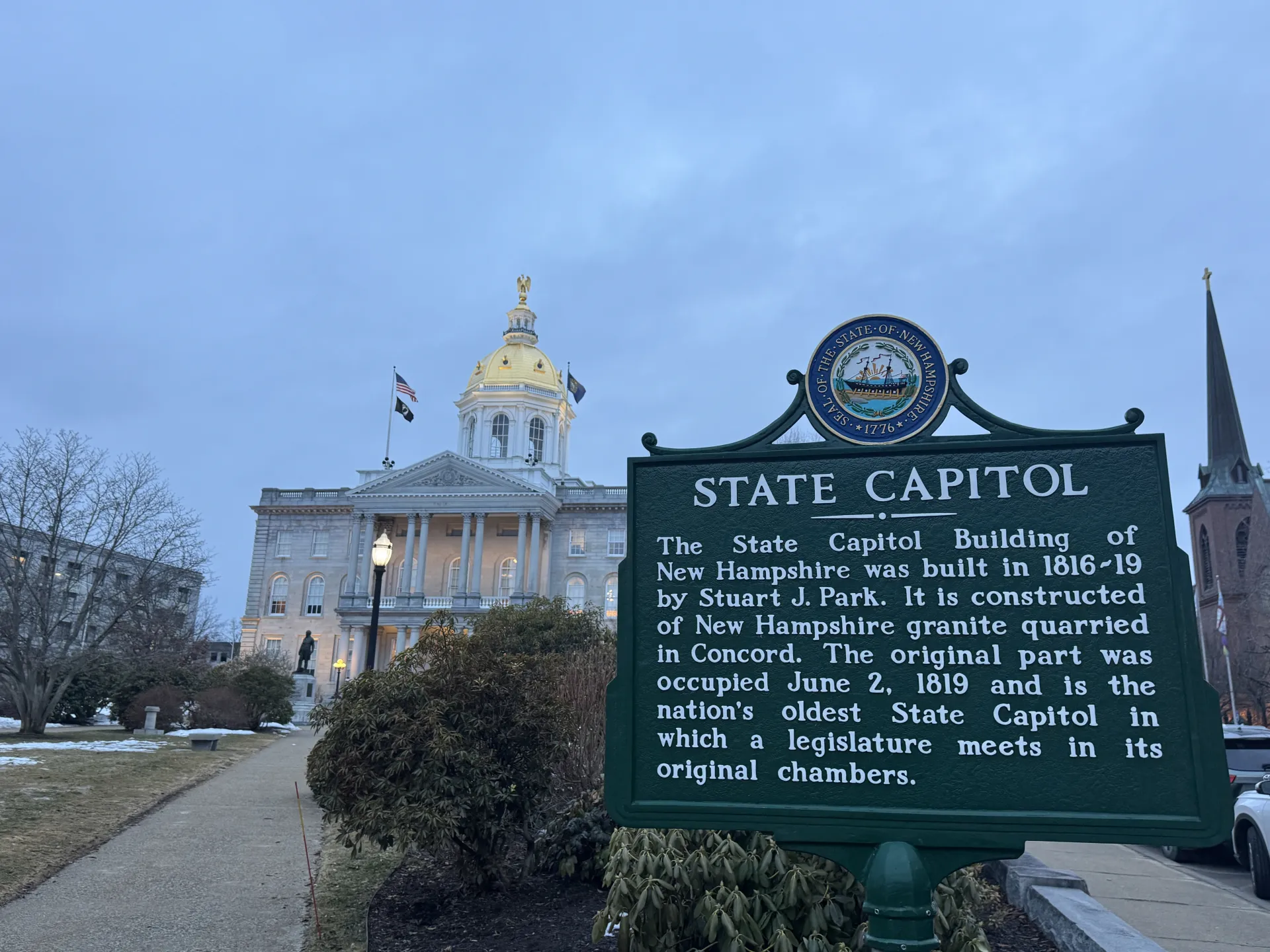 The New Hampshire State Capitol historical sign in the foreground at dusk; the New Hampshire State House in the background.