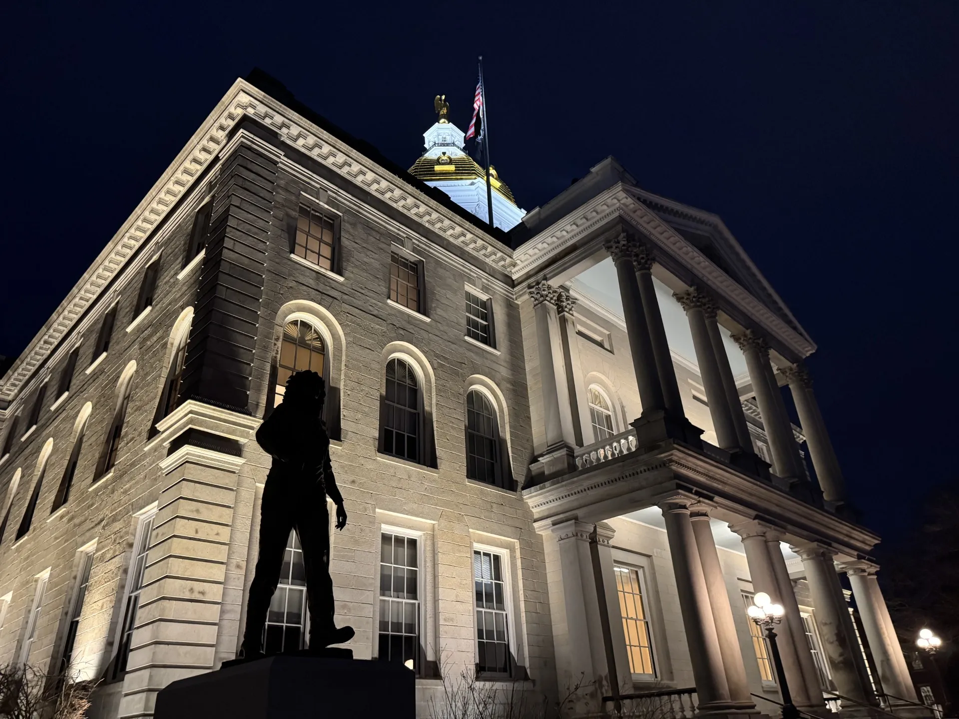 An image of the New Hampshire State House at night, with the Christa McAuliffe statue in the foreground.