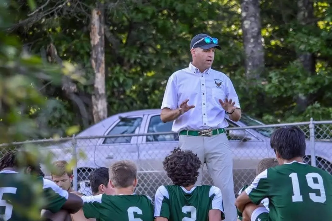 An image of Hopkinton Varsity Boys Soccer Coach Scott Zipke standing before his team who are seated at a game.