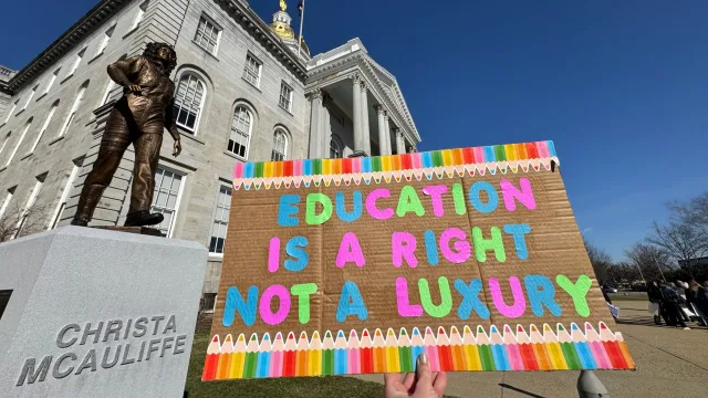 A colorful sign that reads "Education is a luxury not a right." is held in front of the NH statehouse.